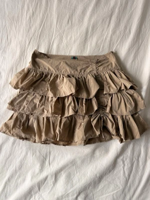 Beige volangkjol från Venezia - Snygg beige kortkjol från Venezia med flera lager volanger som ger en cool och lekfull look. Kjolen har en lätt och luftig känsla, perfekt för varmare dagar. Stängs med dragkedja i sidan.