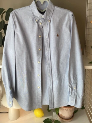 Ljusblå oxfordskjorta från Ralph Lauren - Klassisk ljusblå oxfordskjorta från Ralph Lauren med button-down krage och broderad logga på bröstet. Skjortan har långa ärmar, vita knappar och är tillverkad i ett mjukt bomullstyg. Perfekt för en stilren och avslappnad look.