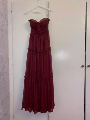 Faeriesty bal-och långklänning  - Romantisk vinröd långklänning med volangdetaljer och axelbandslös design. Klänningen har markerad byst och flera lager i kjolen som ger ett flowy intryck. Perfekt för speciella tillfällen och sommarens fester. Har aldrig använts då jag valde en varmare balklänning. 