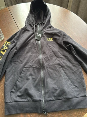 Svart EA7 hoodie med dragkedja - Svart hoodie från EA7 Emporio Armani med gul logga på bröstet och stort tryck längs ena ärmen. Hoodien har dragkedja framtill, två fickor och justerbar huva. Tillverkad i mjuk bomullsblandning för en skön och avslappnad look.
