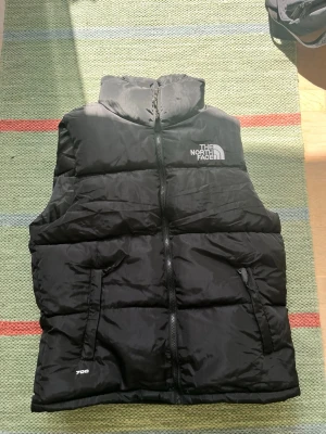 Svart dunväst från The North Face - Säljer en svart dunväst från The North Face med hög krage och dragkedja framtill. Västen har två fickor med dragkedja och logga på bröstet samt 700-brodyr vid nederkanten. Perfekt för lager-på-lager och streetwear.