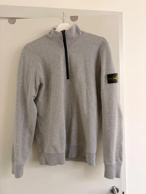 Grå half-zip tröja Stone Island - Snygg grå tröja från Stone Island med half-zip framtill och ikonisk patch på vänster ärm. Tillverkad i mjuk bomullsblandning och har ribbade muddar vid ärmslut och nederkant. Perfekt för en clean och avslappnad stil. Tröjan kostar runt 4000kr nypris och är andvänd ett fåtal gånger.