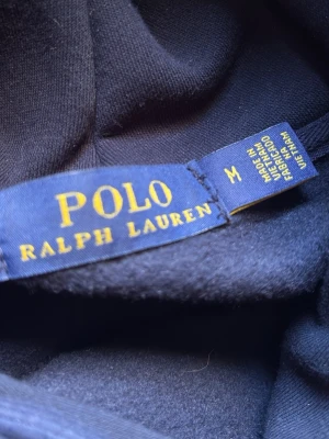 Svart Polo Bear hoodie från Ralph Lauren - Svart hoodie från Polo Ralph Lauren med ikoniska Polo Bear-trycket på bröstet. Klassisk huva med dragsko och mjukt material. Perfekt för dig som gillar streetwear och vill ha en stilren men lekfull look.