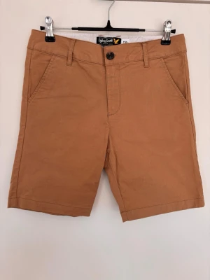 Beiga shorts, Lyle & Scott, 12-13 år - Snygga beiga shorts från Lyle & Scott med klassisk passform. Shortsen har sidofickor, bakfickor med knappar och bälteshällor. Tillverkade i bomull, perfekta för varma dagar. Liten broderad logga bak. Blå shortsen på köpet då det är lite skavt enl bild.