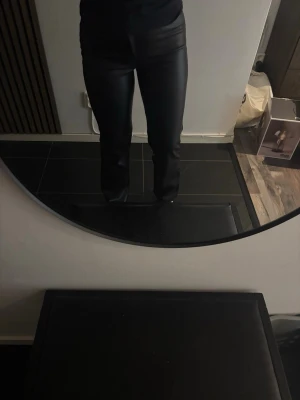 Svarta raka byxor i skinnimitation - Snygga svarta byxor i skinnimitation med raka ben och hög midja. Byxorna har klassisk femficksdesign och stängs med dragkedja och knapp. Perfekta för en trendig och edgy look. Näst intill aldrig använda, otroligt skönt material inte alls stelt men inget som passar mig tyvärr.