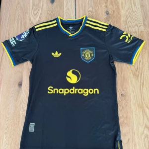 Manchester United svart matchtröja - Svart Manchester United matchtröja från Adidas med gula detaljer och loggor. Tröjan har klubbmärke, Snapdragon-tryck och nummer 37 med namnet Mainoo på ryggen. Korta ärmar och sportig passform, perfekt för fotbollsträning eller support.