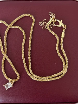 Guldhalsband med rektangulär kristall - Snyggt halsband i guldfärgad metall med en rektangulär klar kristall som hänge. Kedjan har en detaljerad, tvinnad design och justerbar längd med karbinlås. Perfekt för dig som gillar stilrena och glittriga accessoarer.