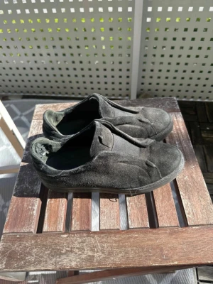 Svarta slip-on sneakers i mocka - Säljer ett par svarta slip-on sneakers i mjuk mocka med minimalistisk design. Skorna har rund tå och svart sula, utan synliga snören eller loggor. Perfekta för dig som gillar enkel och clean stil. Passar 42-42,5