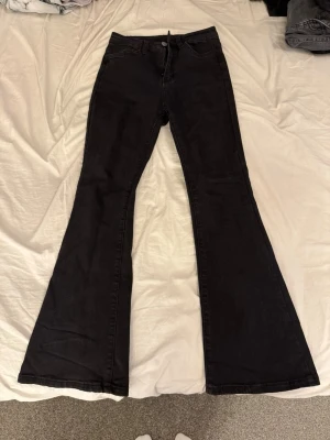 Svarta utsvängda jeans - Säljer ett par svarta jeans med utsvängda ben och klassisk femficksdesign. Jeansen har hög midja och stängs med dragkedja och knapp. 