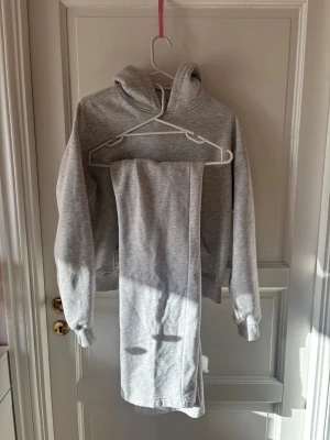 Grått mjukis-set med hoodie,Gina  - Mysigt loungewear-set i ljusgrått med en hoodie och matchande byxor. Hoodien har huva och ribbade muddar vid ärmslut och nederkant. Byxorna är raka och mjuka, perfekta för avslappnade dagar hemma.