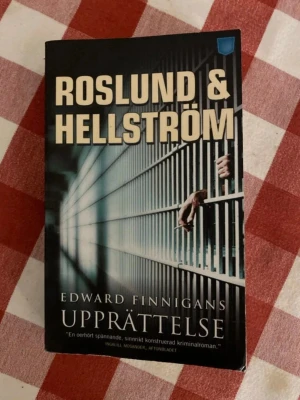 Edward Finnigans upprättelse - Spännande kriminalroman av Roslund & Hellström. Boken handlar om identitet, rättvisa och politiska spel, med Ewert Grens som kriminalkommissarie. Omslaget är svart med fängelsegaller och händer, pocketformat.