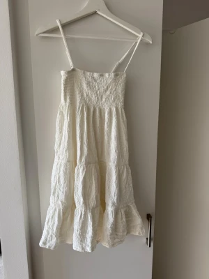 Vit sommarklänning från Gina Tricot - Supersöt vit klänning från Gina Tricot med smock upptill och tunna axelband. Klänningen har volangdetaljer och är gjord i ett lätt, strukturerat tyg som passar perfekt till varma dagar. 