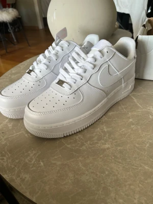 Nike Air Force 1 vita sneakers - Klassiska Nike Air Force 1 sneakers i helvitt läder med perforerad tå, vit sula och vita skosnören. Swoosh-logga på sidorna och metallbricka med 'AF1' på snörningen. Perfekt för en clean och stilren look.