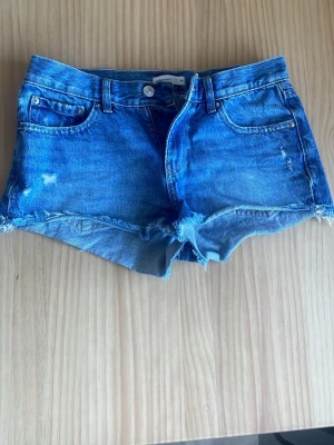 Blå jeansshorts  - Snygga blå jeansshorts från Gina Tricot med fransig kant och slitna detaljer. Klassisk femficksmodell med knapp och dragkedja framtill. Perfekta för varma dagar och en avslappnad stil.