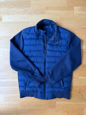 Blå cardigan från Polo Ralph Lauren - Snygg blå cardigan från Polo Ralph Lauren med quiltad framsida och mjuka ärmar i sweatshirtmaterial. Dragkedja framtill, ståkrage med tryckknappar och ribbade muddar. Perfekt för lager-på-lager och streetstyle.