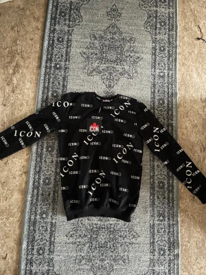 Svart sweatshirt från ICON - Cool svart sweatshirt från ICON med vitt logotyptryck över hela tröjan och en röd detalj på bröstet. Tröjan har rund halsringning och ribbade muddar vid ärmslut och nederkant. Perfekt för en streetwear-look.