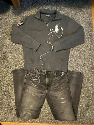 Grå halvzip från Polo Ralph Lauren - Snygg grå tröja med halv dragkedja från Polo Ralph Lauren. Stor vit polospelare broderad på bröstet och patch på ena ärmen. Perfekt för en avslappnad men stilren look.