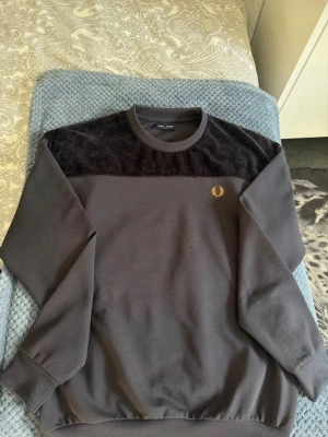 Svart och grå sweatshirt från Fred Perry - Storlek S fast passar nog bäst med M size. Snygg sweatshirt från Fred Perry i mörkgrått med svart sammetsliknande panel upptill. Tröjan har rund halsringning, långa ärmar och den klassiska Fred Perry-loggan broderad i gult på bröstet. Perfekt för en stilren och avslappnad look.