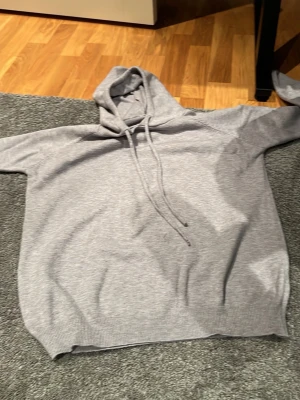 Grå hoodie med dragsko - En stilren grå hoodie med huva och dragsko. Klassisk modell med ribbade muddar i ärmslut och nederkant. Perfekt för en avslappnad look. Materialet känns mjukt och bekvämt, passar till det mesta i garderoben. Tydliga defekter på ena snöret syns änven bara på nära håll!