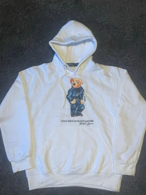 Vit hoodie Polo Bear Ralph Lauren - Vit hoodie från Ralph Lauren med ikoniska Polo Bear-trycket på bröstet. Klassisk huva med dragsko och känguruficka framtill. Mjuk bomullskvalitet och avslappnad passform. Perfekt för en chill och trendig look.