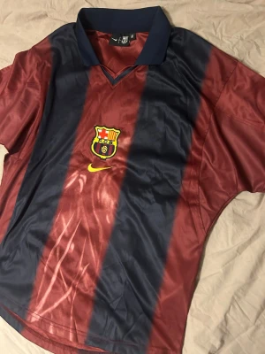 Barça x Cactus Jack matchtröja - Säljer en Barcelona matchtröja i vinrött och marinblått med breda ränder, Nike-logga och klubbmärke på bröstet. Baksidan har tryck med Cactus Jack, nummer 2 och BARÇA. Klassisk krage och korta ärmar. Perfekt för fotbollsfans!