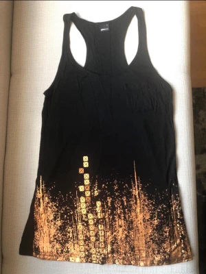 Svart Gina Tricot tank top med roséguldstryck (L) - Säljer en Lilly Tank Top från Gina Tricot i storlek L. Linnet är i en klassisk svart färg med ett effektfullt, glittrigt grafiskt tryck i roséguld nertill. Modellen har en smickrande brottarrygg (racerback) och en liten ficka framtill. Eftersom den är tillverkad i 100% viskos är den supermjuk och har ett lyxigt fall. Sitter snyggt oversized om du brukar bära mindre storlekar! Mycket fint skick.