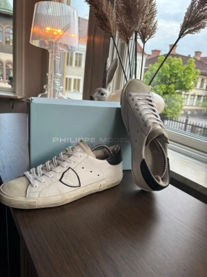 Vita Philippe Model sneakers - Snygga vita sneakers från Philippe Model med svart detalj på hälen och den klassiska sköldloggan på sidan. Skorna har vita snören, låg profil och är tillverkade i läder med en slitstark gummisula. Perfekta för en clean och trendig look.  DOM HAR ENDAST NÅGOT SLITAGE I HÄLKAPPAN, SKRIV FÖR FLER BILDER 