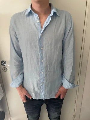 Ljusblå linneskjorta  - Säljer en stilren ljusblå skjorta från Uniqlo i svalt linne. Skjortan har klassisk krage, knäppning framtill och långa ärmar. Perfekt för en avslappnad men snygg look. Passar dig som gillar enkel och fräsch stil. Modellen på bilden är 185 cm och väger 75 kg.
