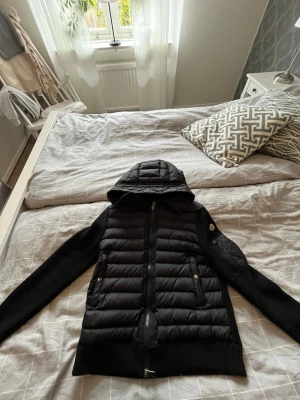 Svart Moncler cardiganjacka med huva - Svart cardiganjacka från Moncler med stickade ärmar och vadderad front. Jackan har huva, dragkedja framtill och två fickor med dragkedja. Moncler-logga på ena ärmen. Perfekt för lager-på-lager och snygg streetstil.