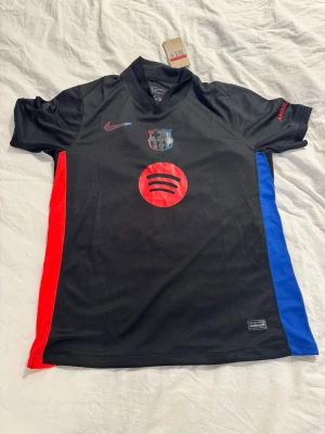 FC Barcelona svart matchtröja Pedri - Svart FC Barcelona matchtröja från Nike med röda och blå detaljer på sidorna. Tryck med Spotify-logga framtill och Pedri 8 på ryggen. Klubbmärke och Nike-logga på bröstet. Korta ärmar och sportig passform, perfekt för fotbollsträning eller match.