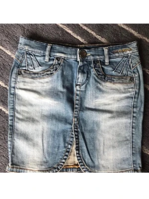 Blå jeanskjol från Killah - Snygg ljusblå jeanskjol  med klassisk femficksdesign och dekorativa sömmar vid fickorna. Kjolen har bälteshällor, knapp och dragkedja framtill samt en liten slits. Perfekt för dig som gillar en avslappnad och trendig stil.