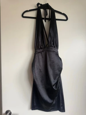 Svart halterneck klänning  - Snygg svart kort klänning  med halterneck och öppen rygg. Klänningen har en tight passform och knyts i nacken för en cool look. Perfekt för dig som vill sticka ut med en elegant och stilren vibe.