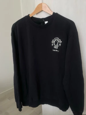 Svart Keith Haring HM sweatshirt - Svart sweatshirt med ikoniskt Keith Haring-tryck i vitt på bröstet och stort grafiskt motiv på ryggen. Tröjan har rund halsringning och långa ärmar. Tillverkad i mjuk bomull för en skön och avslappnad look.