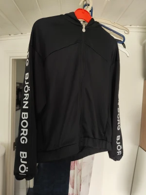 Svart hoodie från Björn Borg - Svart hoodie från Björn Borg med dragkedja framtill och huva. Stora vita logotexter längs båda ärmarna ger en sportig vibe. Tillverkad i ett mjukt och stretchigt material som känns skönt mot huden. Perfekt för dig som gillar streetwear och vill ha en clean look.