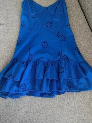  Nå trendig Blå kort klänning med volang - Snygg blå kort klänning med smala axelband och volangkant nertill. Klänningen har broderade blommönster och är tillverkad i ett lätt bomullstyg. Perfekt för varma dagar och har en feminin, luftig känsla.
