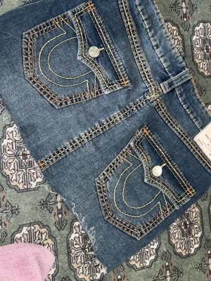 True Religion jeanskjol med stora fickor - Säljer en blå jeanskjol från True Religion med stora bakfickor, kontrastsömmar och rå kant nertill. Kjolen har klassiska silverknappar och tydliga gula och orange sömmar som ger en cool streetkänsla.