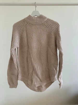 Beige stickad tröja från Gina Tricot - Säljer en beige stickad tröja från Gina Tricot med hög krage och grovstickat mönster. Tröjan har lång ärm, ribbade muddar och en något längre bakdel. Perfekt för lager-på-lager och chill dagar.