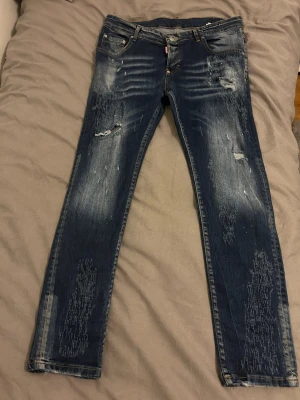 DESQUARED2 blå jeans - Säljer ett par mörkblå jeans med raka ben och tydliga slitningar samt distressed detaljer framtill och baktill. Jeansen har klassisk femficksdesign och en grön patch bak i midjan. Perfekta för en avslappnad och trendig look. Desquard2 jeans