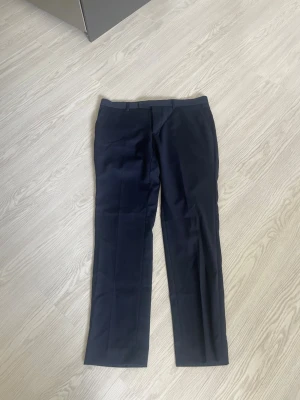 Mörkblå kostymbyxor Dressmann slim fit - Snygga mörkblå kostymbyxor från Dressmann i slim fit-modell. Byxorna har klassisk design med pressveck, bälteshällor och raka ben. Perfekta för en stilren look. Materialet känns mjukt och följsamt.