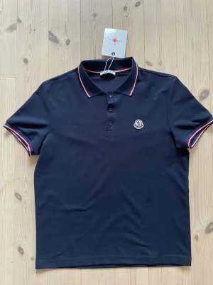 Mörkblå piké  - Klassisk mörkblå piké med korta ärmar och diskret logotyp på bröstet. Toppens krage och ärmslut har snygga rödvita detaljer. Passa nog bäst mellan 175-180 cm beroende på vikt och kroppstyp. 