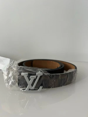 Louis Vuitton brunt bälte med LV-spänne - Snyggt bälte från Louis Vuitton i brunt skinn med klassiskt LV-spänne i silverfärg. Bältet har ett diskret rutigt mönster och insida i ljusare beige ton. Perfekt accessoar för att lyfta din outfit. Köpt utomlands.