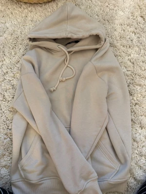 Beige hoodie från BikBok - Snygg beige hoodie från BikBok med klassisk huva och dragsko. Hoodien har en stor magficka och ribbade muddar vid ärmslut och nederkant. Perfekt för en chill och avslappnad stil. Tillverkad i mjukt bomullsmaterial.
