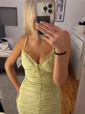 Ljusgul kort klänning med smala axelband - Säljer en ljusgul kort klänning med smala axelband och rynkade detaljer framtill. Klänningen har en figurnära passform och subtilt blommigt mönster. Perfekt för varma dagar och somriga tillfällen.