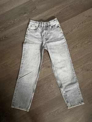 Grå jeans med hög midja - Säljer ett par grå mom jeans med hög midja och raka ben. Jeansen har klassisk femficksdesign och knappgylf. Materialet är kraftig denim i tvättad look som ger en retrovibe. Perfekta för dig som gillar avslappnad men trendig stil.