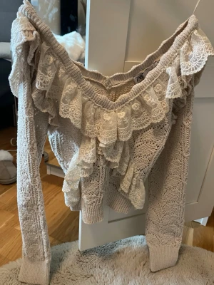Beige stickad tröja med spetsvolanger - Supersöt beige stickad tröja från Zara med breda spetsvolanger över axlar och bröst. Tröjan har lång ärm, v-ringad hals och är gjord i ett mjukt stickat material med detaljerad spets för en romantisk look.