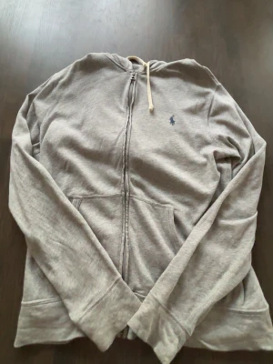 Grå hoodie från Polo Ralph Lauren - Snygg grå hoodie med dragkedja från Polo Ralph Lauren. Klassisk design med huva och dragsko, två fickor framtill och den ikoniska lilla loggan broderad på bröstet. Perfekt för en chill och avslappnad stil.