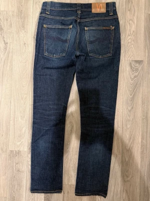 Nudie Jeans Grim Tim Dry Jeans Mörkblå W32/L32 - Slim Straight Fit - Nudie Grim Tim mörkblå jeans i organic dry denim med slim straight fit passform som sitter snyggt men mer rakt i benet och funkar till allt, storlek W32/L32, midja 39 cm, ytterbenslängd 100,5 cm, benöppning 17 cm, modellen är 185cm & väger 65kg, i fint skick.