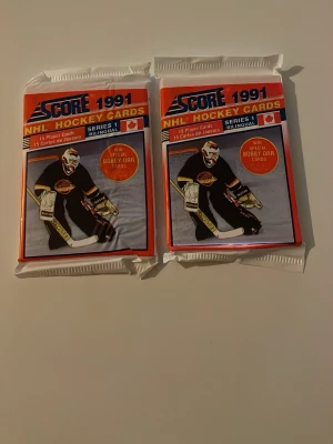 Score 1991 NHL hockeykort, 2 oöppnade - Två oöppnade paket Score 1991 NHL hockeykort, Series 1. Varje paket innehåller 15 spelarkort och är tvåspråkiga. Framsidan visar en målvakt i svart matchställ. Perfekt för samlare eller hockeyfans!