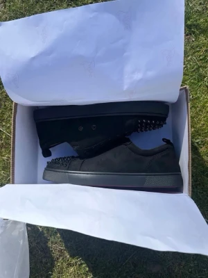 Svarta sneakers med nitar Christian Louboutin - Svarta sneakers i mocka och läder från Christian Louboutin med coola svarta nitar på tån. Skorna har lågt skaft, svarta skosnören och en klassisk röd sula. Perfekta för dig som vill sticka ut med en edgy stil.