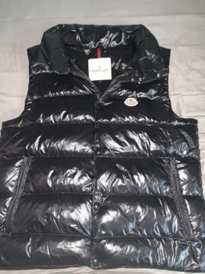 Svart dunväst från Moncler - Snygg svart dunväst från Moncler med glansig finish och klassisk logga på bröstet. Västen har dragkedja framtill, två fickor med dragkedja och hög krage. Perfekt för lager-på-lager och streetstyle.